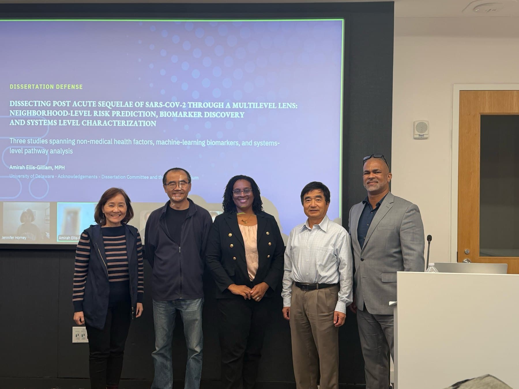 Five people standing in a row in front of a projected title slide reading “Dissertation Defense: Dissecting Post Acute Sequelae of SARS-CoV-2 Through a Multilevel Lens.” Amirah Ellis-Gilliam stands in the middle in a dark blazer, flanked by her committee members; a Zoom panel on the screen shows Jennifer Horney attending remotely.