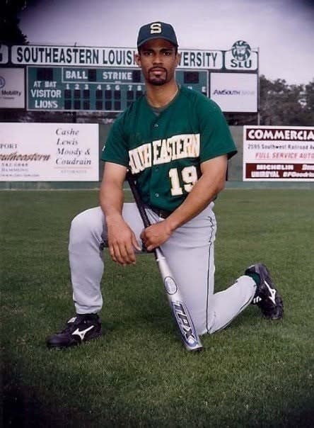 Félix E. Rivera-Mariani kneeling on the outfield grass in a Southeastern Louisiana University baseball uniform, number 19, holding a bat in front of the stadium scoreboard.